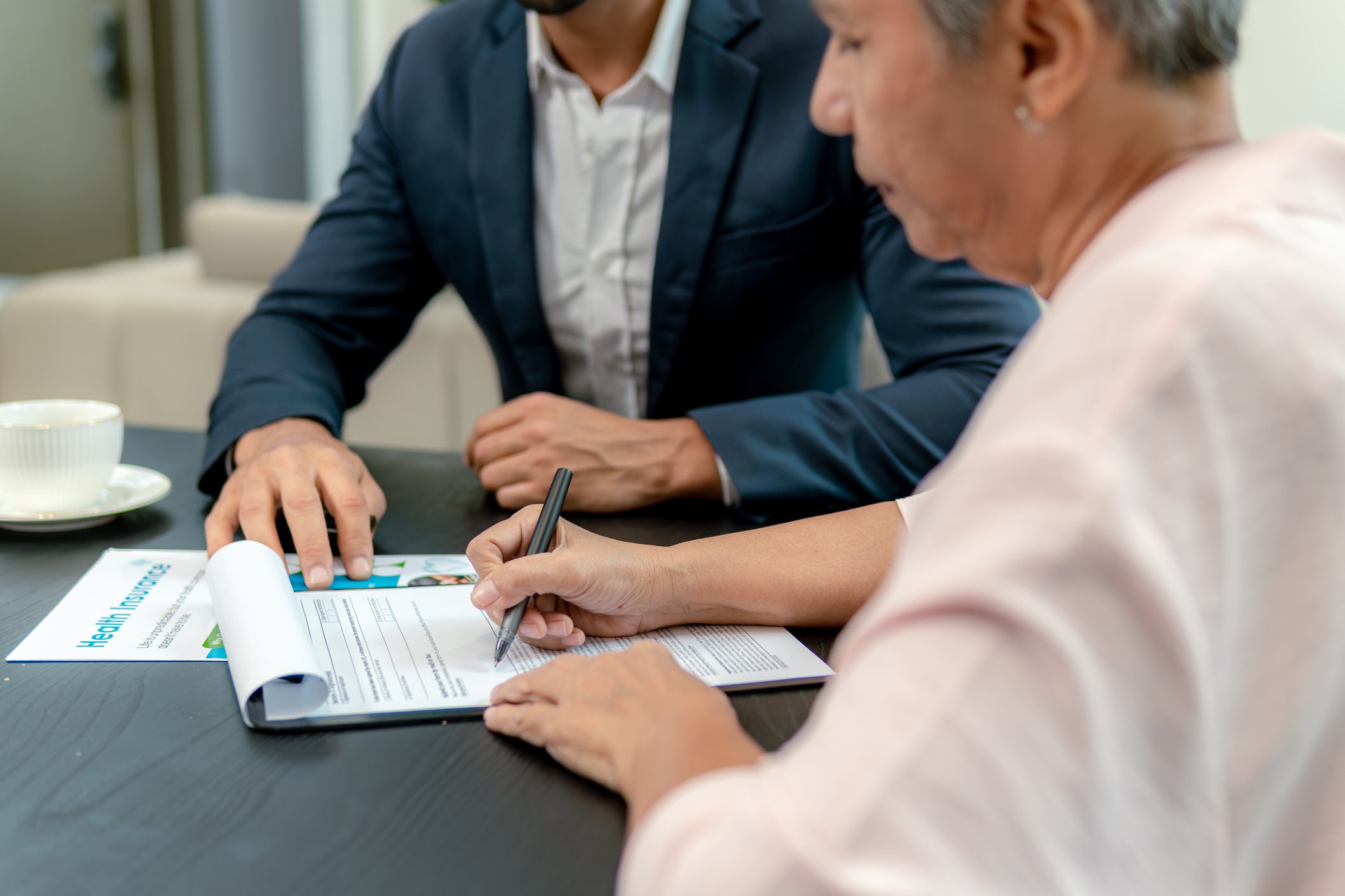 An older woman signs a Medicare Advantage plan application while consulting with a professional at a table.