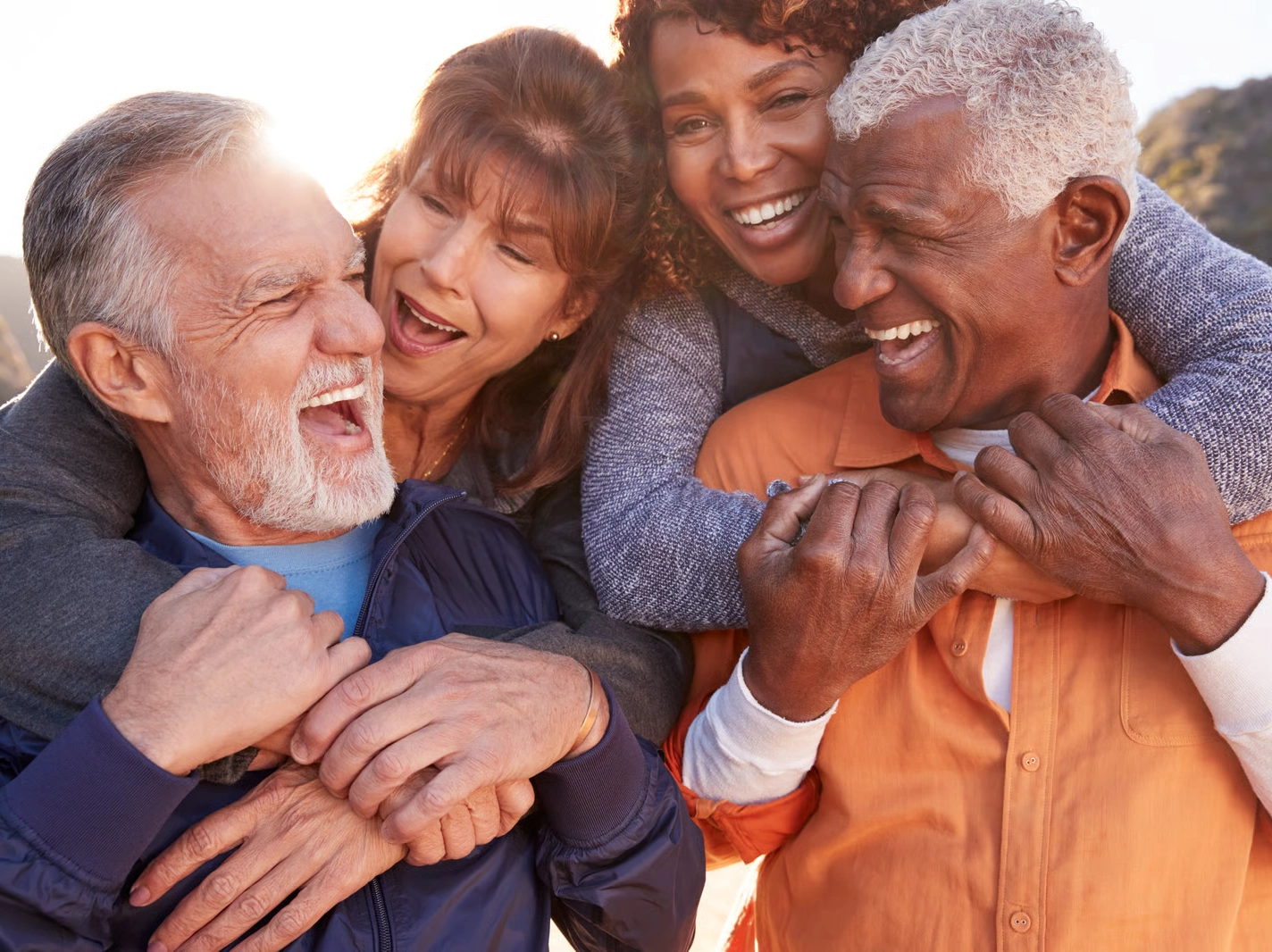 Four diverse seniors joyfully embrace and laugh together outdoors, symbolizing community and connection in retirement.