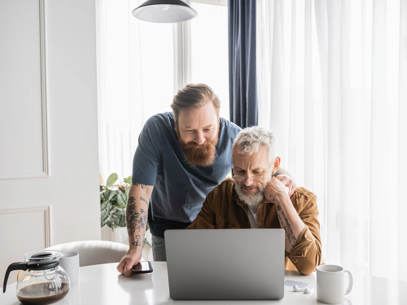 Two men, one older and one younger, review Medicare enrollment information on a laptop at a bright, home setting.