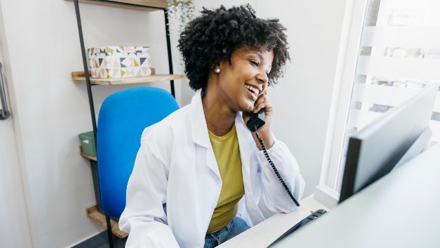 A healthcare professional in a lab coat smiles while speaking on the phone at a desk with a computer.