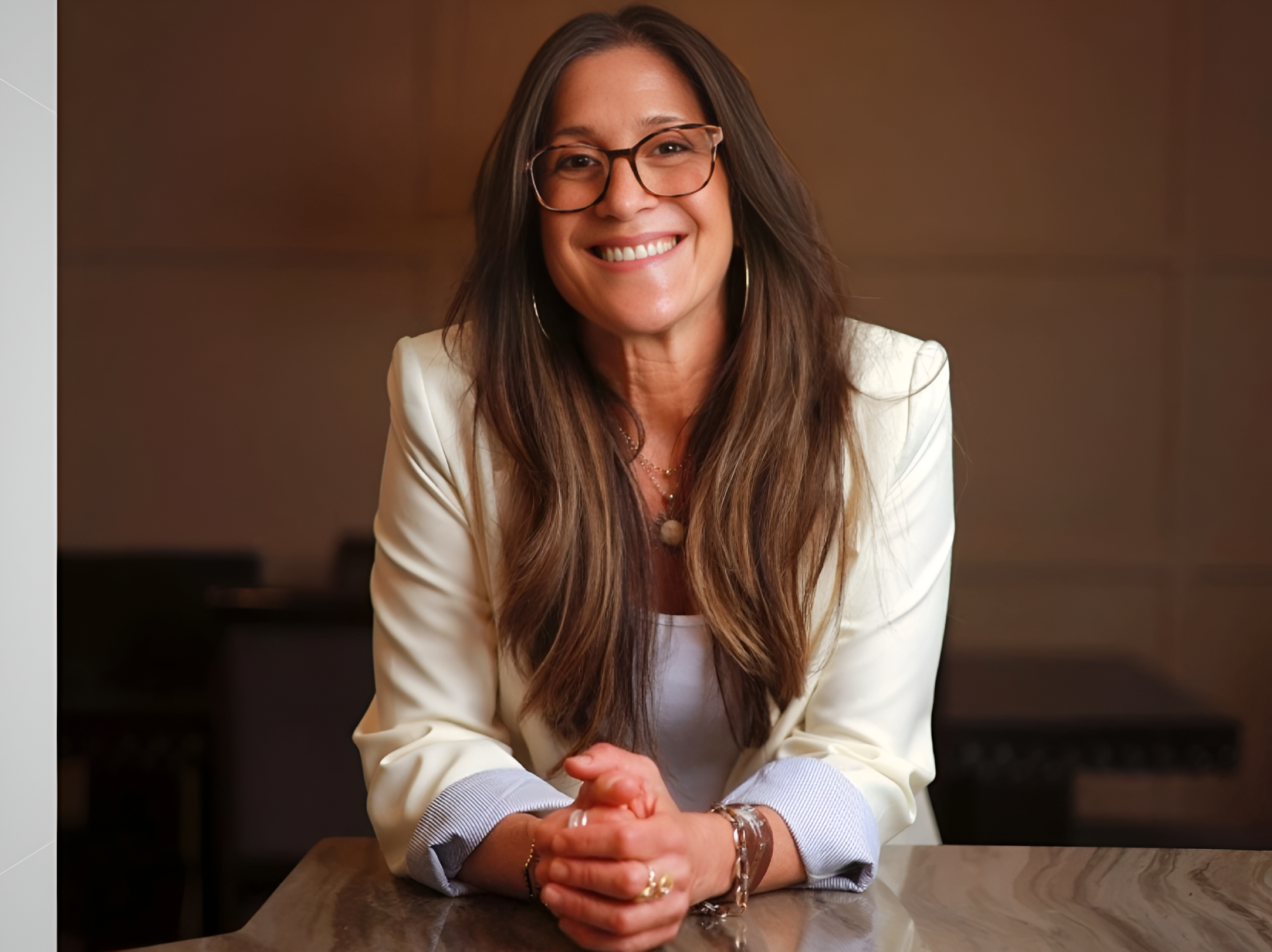 Smiling woman with long hair and glasses leans on a table, dressed in a white blazer, conveying approachability and professionalism.