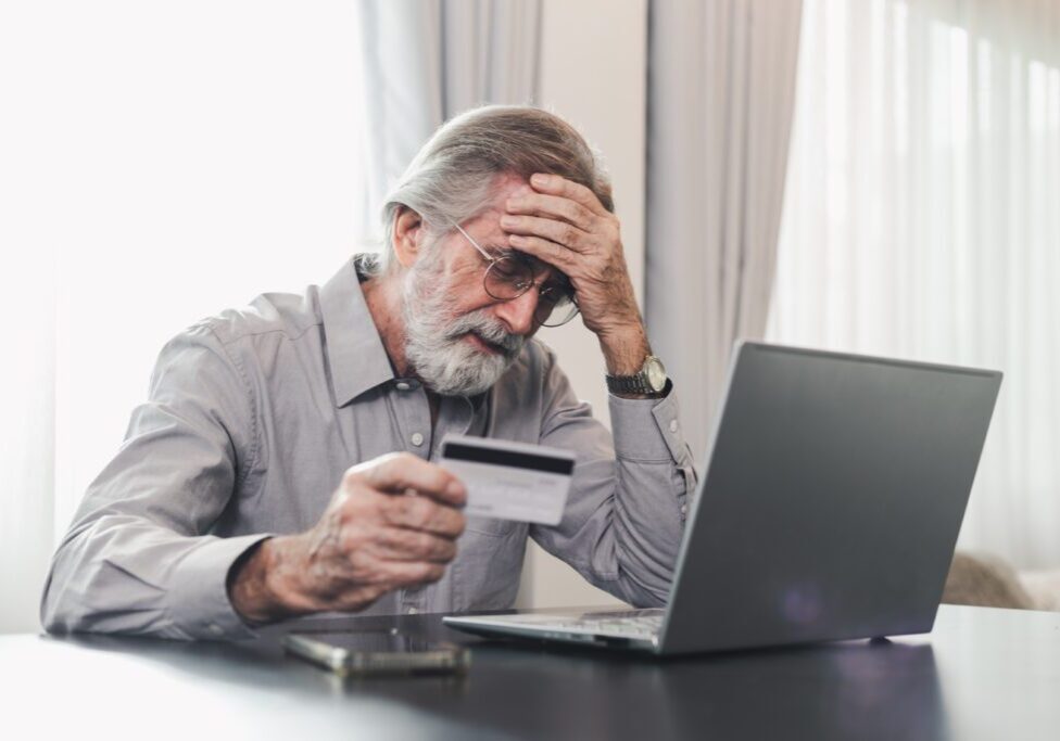 Upset senior elderly man holding credit card by laptop having trouble worry finance safety data or online payment security. Bank client concerned about problem with credit card, financial fraud threat